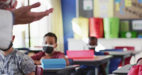 Diverse teacher disinfecting hands in classroom, with schoolchildren ...