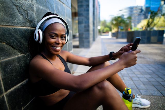 Smiling fit african american woman wearing headphones, resting, using smartphone in street - Download Free Stock Photos Pikwizard.com