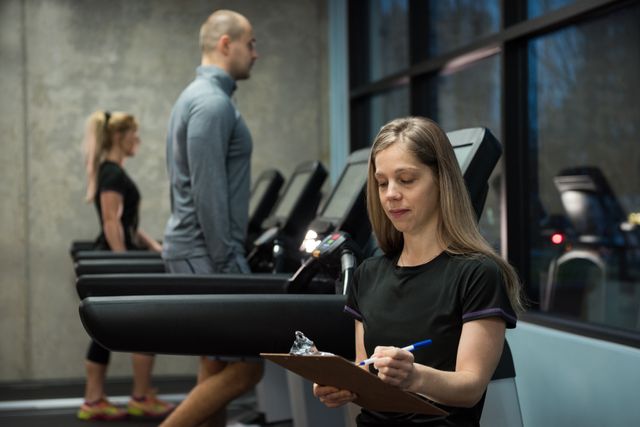 Woman writing on clipboard with people exercising on treadmill - Download Free Stock Photos Pikwizard.com