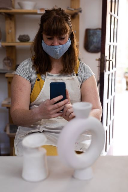 Caucasian female potter wearing face mask using a smartphone - Download Free Stock Photos Pikwizard.com