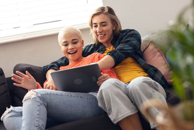 Low angle view of multiracial happy lesbian young couple using digital tablet while sitting on sofa. Wireless technology, unaltered, love, togetherness, homosexual, lifestyle and home concept.