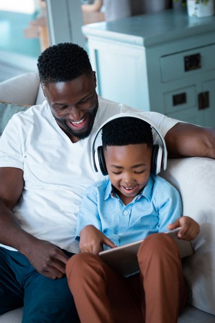 African american father and son sitting on sofa, wearing headphones and using tablet - Download Free Stock Photos Pikwizard.com