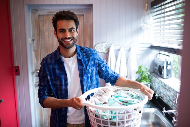 Portrait of smiling young man holding laundry basket while standing by window at home