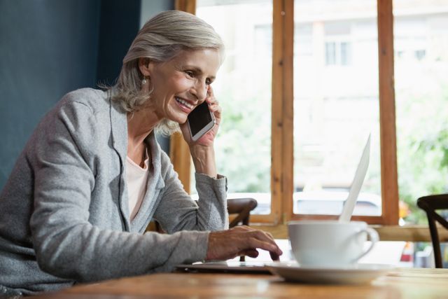 Smiling senior woman talking on mobile phone while working on laptop - Download Free Stock Photos Pikwizard.com
