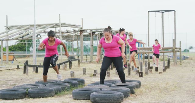 Determined caucasian female friends in pink t shirts crossing logs and tyres at bootcamp training. Female fitness, friendship, challenge and healthy lifestyle.