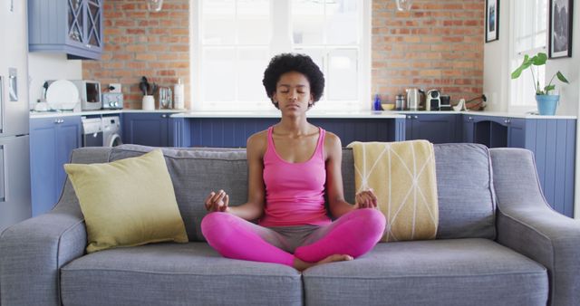 Relaxed african american teenage girl sitting on sofa doing yoga and meditating. domestic life, spending time at home.