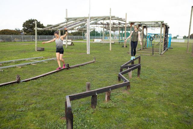 Front view of two Caucasian women wearing sports clothes balancing walking along wooden beams at an outdoor gym during a bootcamp training session, with outdorr gym equipment in the background