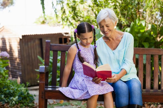 Smiling grandmother reading book to granddaughter while sitting on wooden bench - Download Free Stock Photos Pikwizard.com