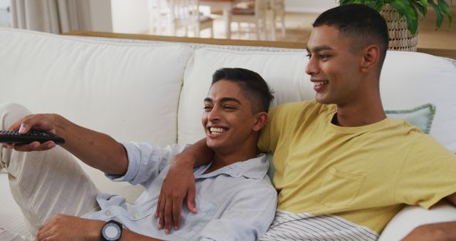 Smiling mixed race gay male couple sitting on sofa watching tv one using remote - Download Free Stock Photos Pikwizard.com