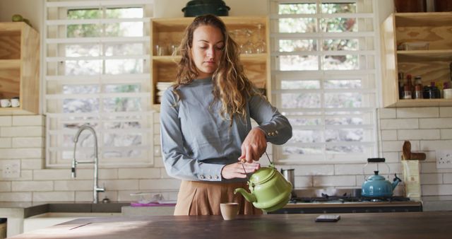 Happy caucasian woman standing at counter in cottage kitchen pouring tea from teapot and smiling. simple living in an off the grid rural home.