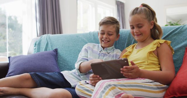 Happy caucasian brother and sister at home, sitting on couch using tablet together and smiling. happy family, at home together.