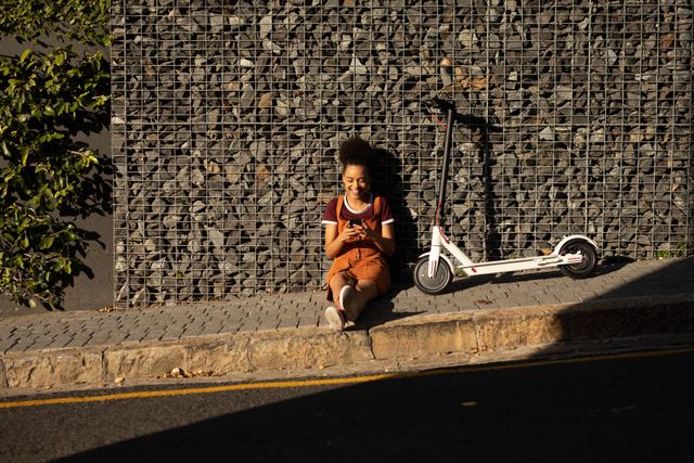 Mixed race woman looking at her phone sitting on the sidewalk - Download Free Stock Photos Pikwizard.com