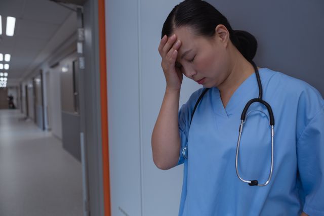 Sad female doctor with hand on forehead leaning against a wall in corridor at hospital - Download Free Stock Photos Pikwizard.com