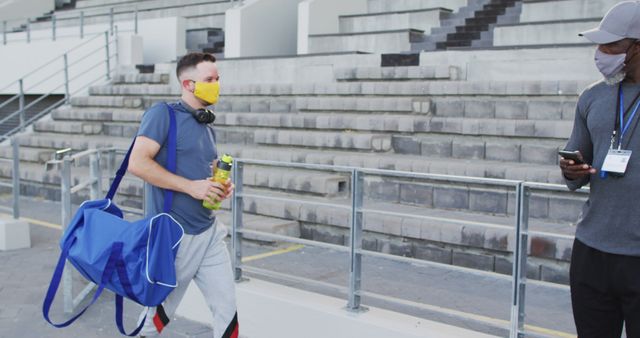 Diverse male coach and disabled athlete wearing face mask touching elbows before training session. professional runner training at sports stadium during coronavirus covid 19 pandemic.