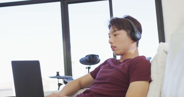 Happy asian male teenager wearing headphones and using laptop in living room. spending time alone at home.