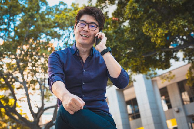 Asian man smiling while talking on smartphone sitting at corporate park - Download Free Stock Photos Pikwizard.com