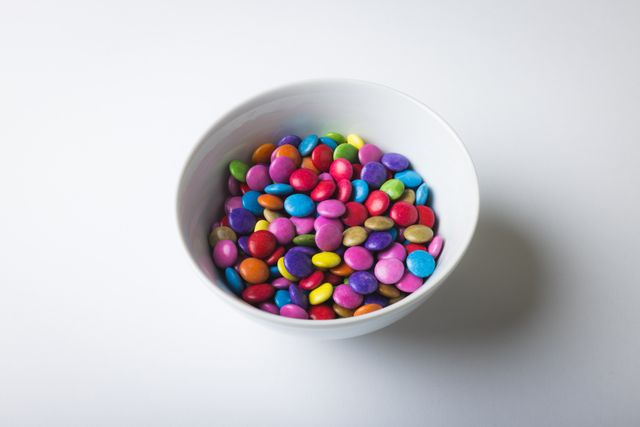 High angle view of multi colored chocolate candies in bowl amidst copy space on white background. unaltered, unhealthy eating and sweet food concept.