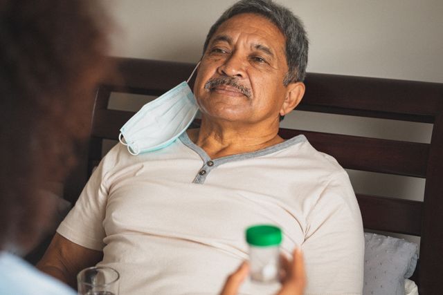 Senior mixed race man wearing face mask lying in bed smiling during medical home visit - Download Free Stock Photos Pikwizard.com