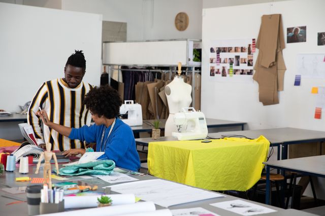 Side view of a young African American male fashion student and a young mixed race female fashion student discussing a design together at a worktable in a studio at fashion college, with mannequins in the background.