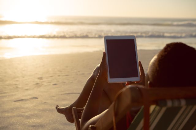 Rear view of woman using digital tablet while relaxing in a beach chair on the beach