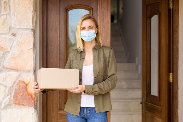 Caucasian woman wearing a mask standing outside the front door of her house while holding a package - Download Free Stock Photos Pikwizard.com