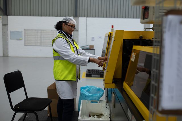 Side view of focused Caucasian female worker working in a factory warehouse, wearing hair net, glasses, lab coat and a high visibility vest, operating a machine on the production line