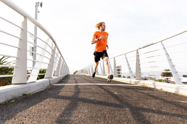 Low angle view of a fit Caucasian man with long blonde hair wearing sportswear exercising outdoors in the city on a sunny day with blue sky, running on a footbridge.