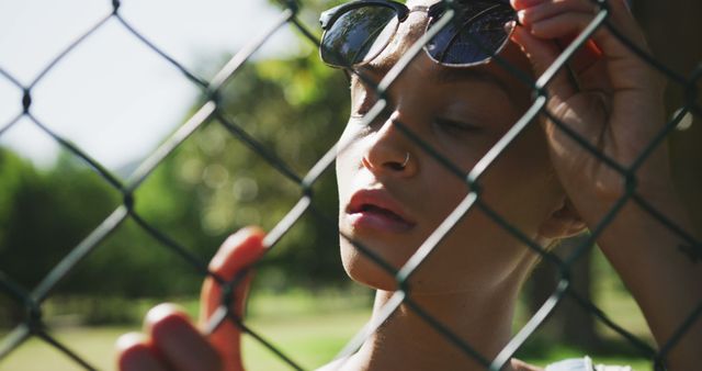 Biracial woman wearing sunglasses and holding chain-link fence at park. Street style and modern urban lifestyle.