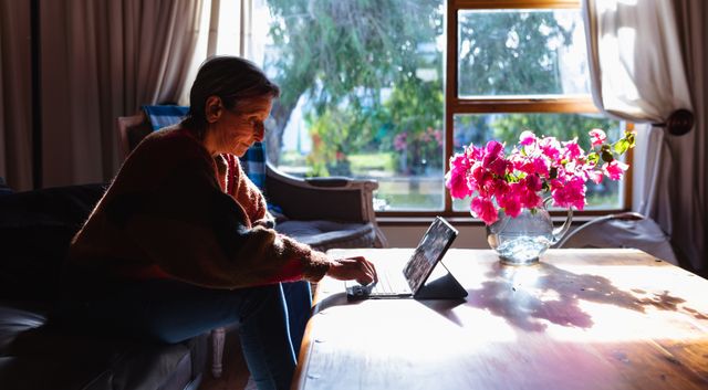 Side view of a senior Caucasian woman with short grey hair wearing a brown sweater sitting on a sofa in her sitting room, using computer laptop, vase with flowers on the table.