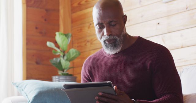 Happy senior african american man spending time in log cabin, sitting on sofa and using tablet. Free time, domestic life and nature concept.