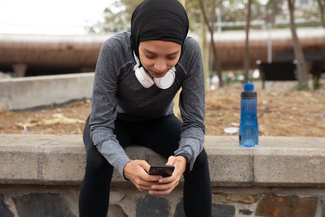 Fit mixed race woman wearing hijab and sportswear exercising outdoors in the city, taking break using smartphone with headphones around her neck. Urban lifestyle exercise.