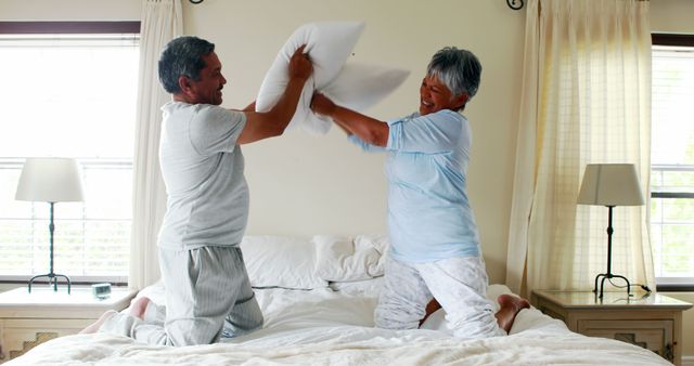 A middle-aged Asian couple is playfully engaging in a pillow fight on a bed, with copy space - Download Free Stock Photos Pikwizard.com