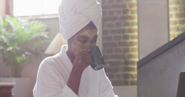 African american attractive woman with applied face mask drinking coffee and using laptop at home. beauty, pampering, home spa and wellbeing concept.
