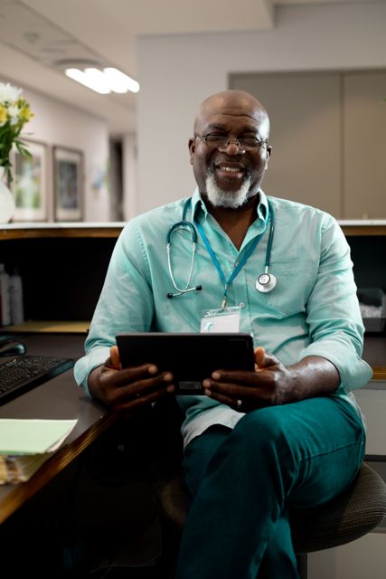 Portrait of smiling senior african american male doctor sitting in hospital reception holding tablet. Medical services, hospital and healthcare concept.