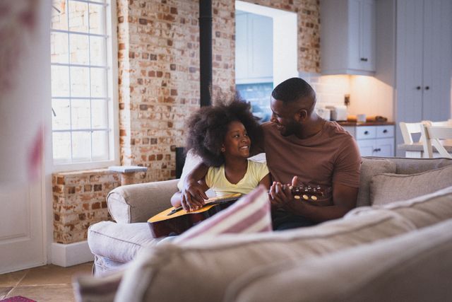 Happy african american girl and her father sitting on couch playing guitar together in living room - Download Free Stock Photos Pikwizard.com