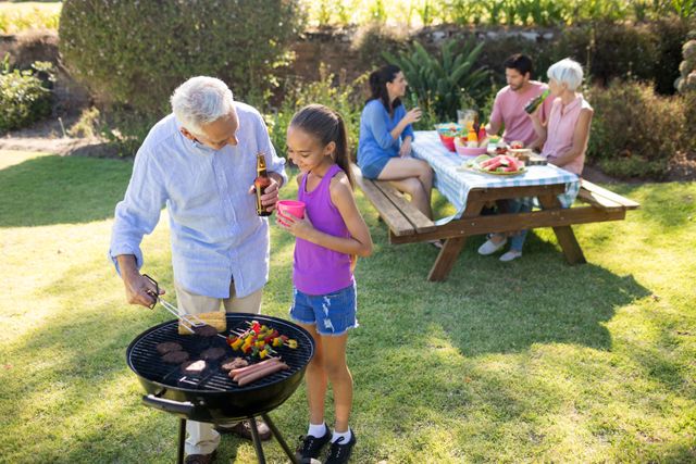 Grandfather and granddaughter preparing barbecue while family having meal in background