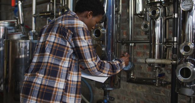 African american woman working at gin distillery checking equipment and writing on clipboard. work at an independent craft gin distillery business.