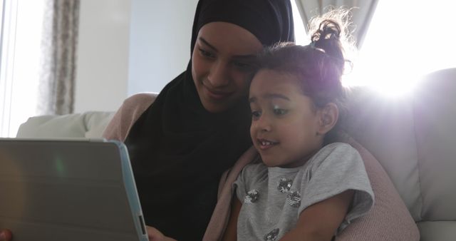 Front view of a young mixed race woman wearing hijab with her young daughter in the sitting room, sitting on a sofa and using a tablet computer