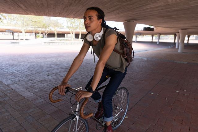 Mixed race alternative man with dreadlocks and headphones out and about in the city on a sunny day, riding bike under bridge. Urban trendy man on the go.