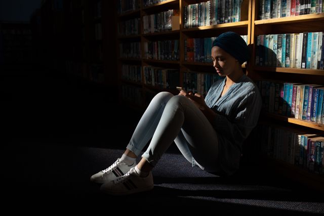 Side view of a mixed race female student wearing a dark blue hijab studying in a library, sitting and holding a book in hands, reading it.