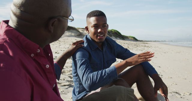 African american father sitting on beach with teenage son, putting hand on his shoulder and talking. healthy outdoor family leisure time together.