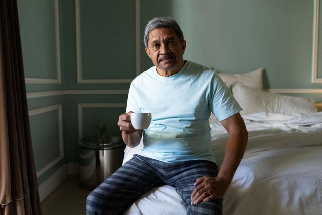 Senior african american man sitting on a bed drinking coffee in a sleeping room - Download Free Stock Photos Pikwizard.com