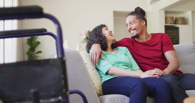 Happy biracial couple sitting on couch embracing in living room, with wheelchair in foreground. wellbeing and domestic lifestyle with physical disability.