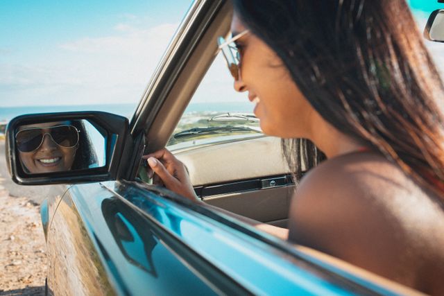 Mixed race woman smiling wearing sun glasses looking in wing mirror in convertible car. summer holiday road trip on a country highway by the coast.