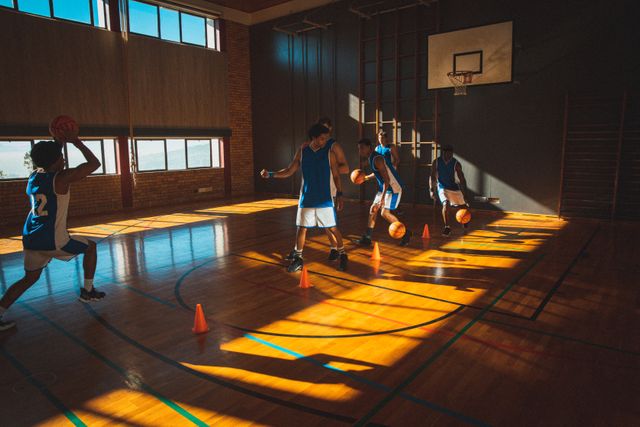 Diverse male basketball team practicing dribbling balls between cones in sunny gym. basketball, team sports training at an indoor court.