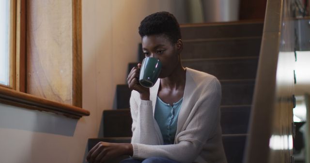 Thoughtful african american attractive woman sitting on stairs an, drinking coffee. leisure time, wellbeing and relaxing at home.