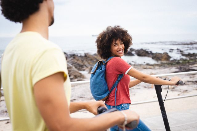 Cheerful afro african american young woman looking at boyfriend while riding push scooter by beach. unaltered, lifestyle, love and togetherness concept.