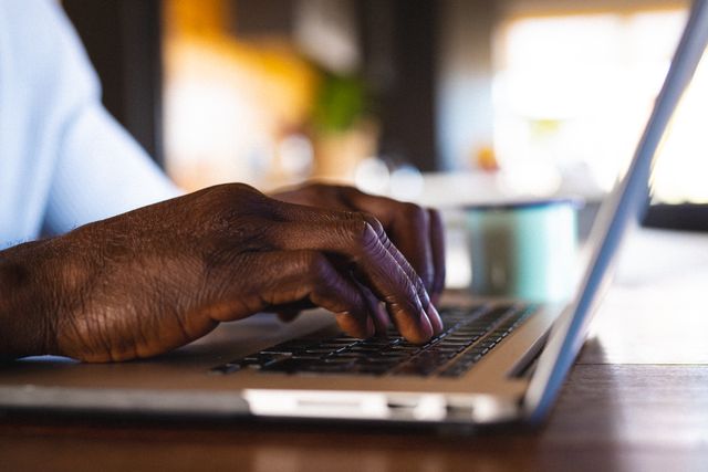 Cropped hands of african american senior man using laptop on table in log cabin. Unaltered, retirement, vacation, solitude, technology, close-up, keyboard, internet and leisure concept.