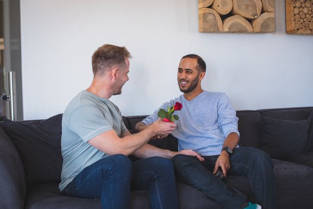 Diverse gay male couple sitting on sofa one giving red rose to the other. staying at home in isolation during quarantine lockdown.