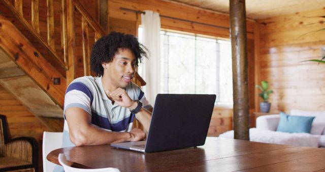 Happy african american man sitting at table and using laptop for image call, slow motion. Lifestyle, domestic life, countryside and nature concept.